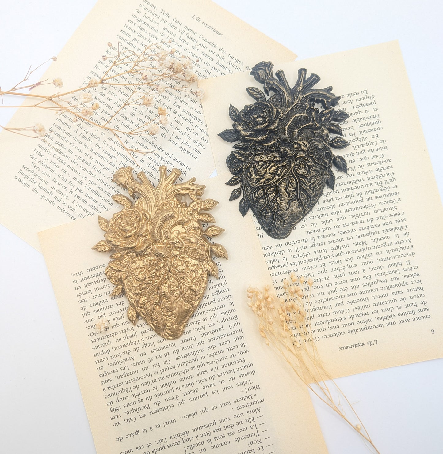 Deux ornements décoratifs en forme de cœur, l'un doré, l'autre bronze foncé. Ils présentent des motifs floraux et sont posés sur des pages de livres anciens avec des fleurs séchées.