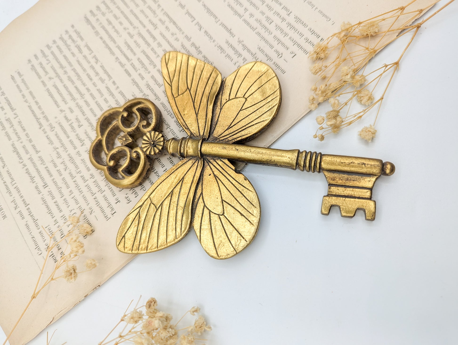 Une clé décorative en laiton avec des ailes de papillon. La clé présente un dessus en forme de nuage et une extrémité en trou de serrure. Les ailes de papillon sont détaillées avec des motifs de veines. La clé mesure environ 15 cm de long.