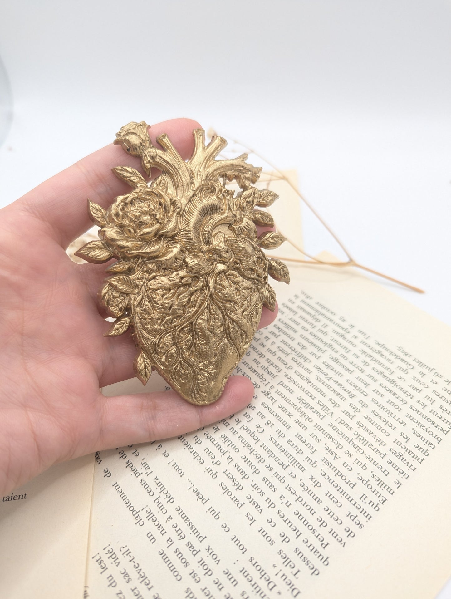 Ornement décoratif doré en forme de cœur avec des accents floraux, posé sur une page de livre vintage. Le cœur présente des détails complexes, probablement un moulage ou un embellissement.